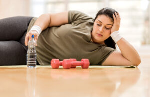 An exhausted woman lying on her side on a workout mat, with a water bottle within reach and a set of dumbbells nearby, appears to be taking a break from exercising.