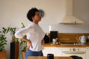 A person standing in a kitchen, facing away from the camera, holding their lower back in a stretch with a pained expression.