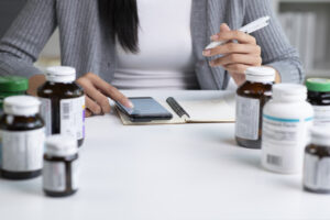 A person at a desk with multiple medicine bottles, pointing at a smartphone lying on a notebook, possibly managing medication or health information, with a pen in hand, wearing a white shirt and grey cardigan.