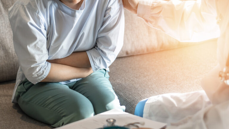 A person sitting on a couch, holding their stomach in discomfort, with a doctor nearby offering support and medical consultation