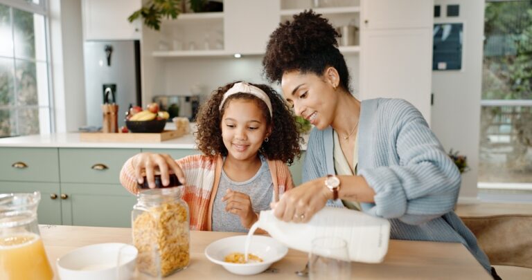 A mother smiles as she pours milk into a cereal bowl for her younger daughter.
