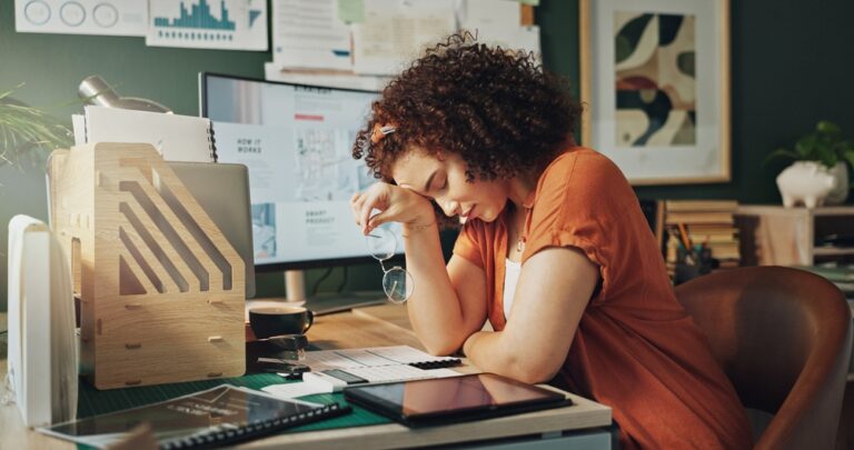 A woman sitting at a cluttered desk in an office, looking stressed and tired. She is resting her head on one hand while holding her eyeglasses in the other. A computer screen, paperwork, tablet, and office supplies surround her. The workspace is decorated with charts, graphs, and framed art on the wall.